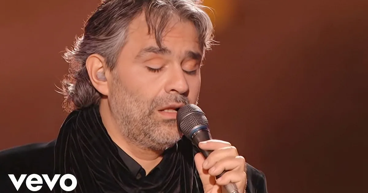 A man with closed eyes sings into a microphone, wearing a black shirt and holding the mic with his right hand.