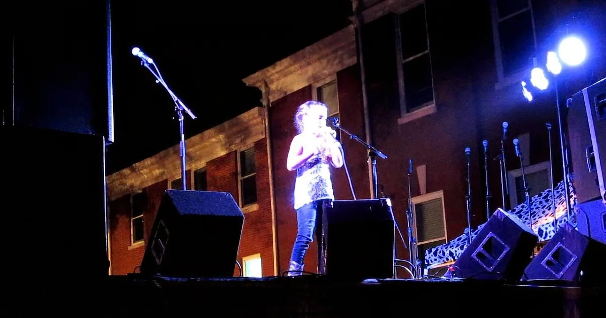 A person stands on a stage with microphones, surrounded by speakers and lights, in front of a brick building at night.