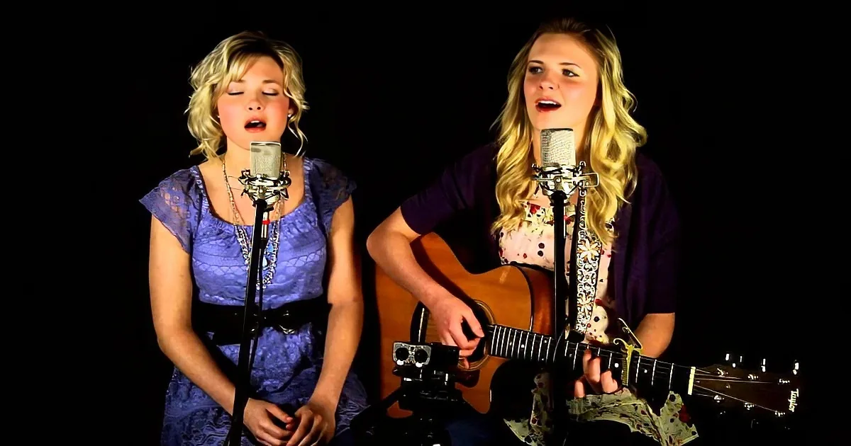 Two women sing into microphones, one plays guitar, both are in front of black background.