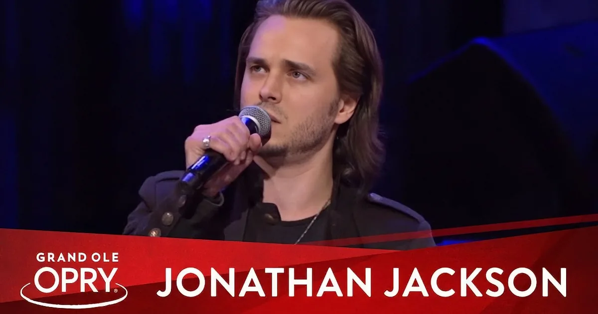 A man with long hair sings into a microphone at the Grand Ole Opry, wearing a black jacket and a necklace.