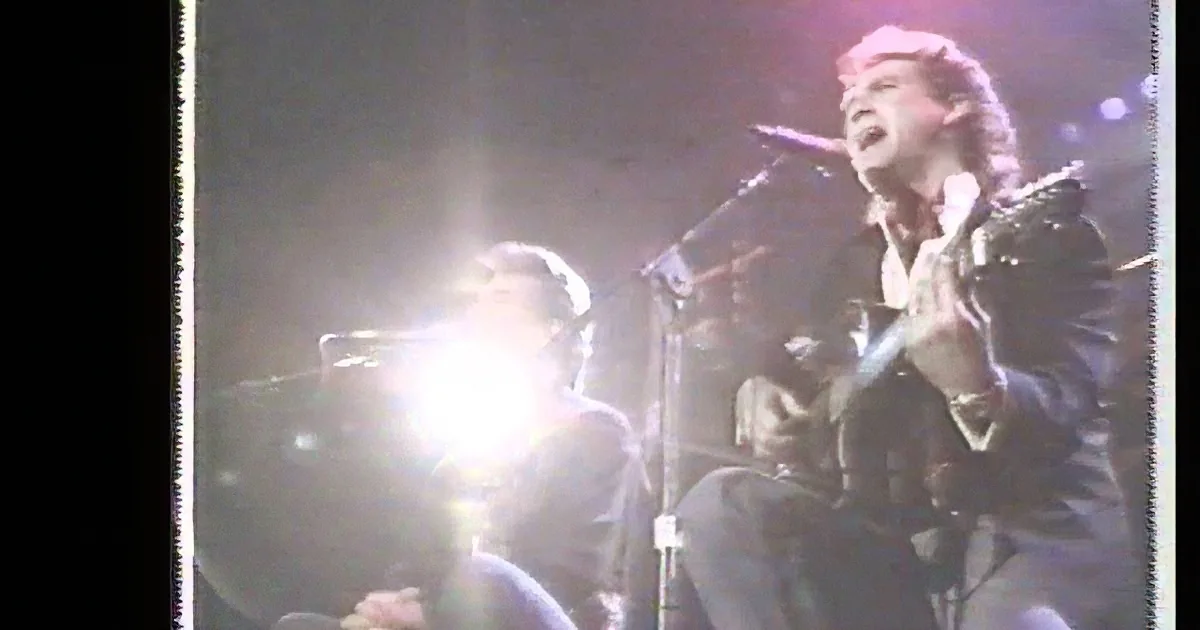 A person in a leather jacket sings into a microphone on stage, illuminated by bright stage lights.