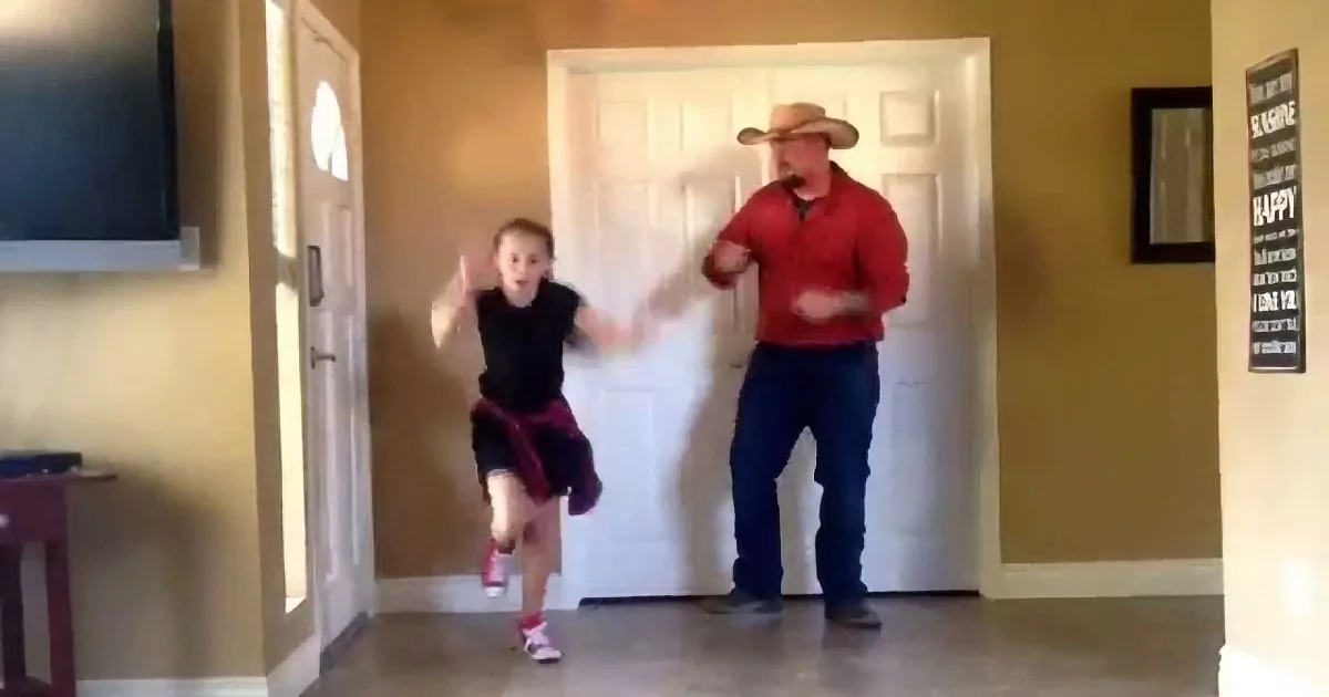 A young girl and an adult in a cowboy hat are dancing in a hallway.
