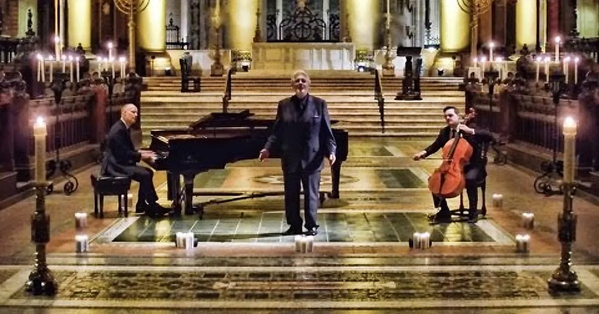 A man sings on stage with a pianist and cellist, surrounded by candles in a grand hall.