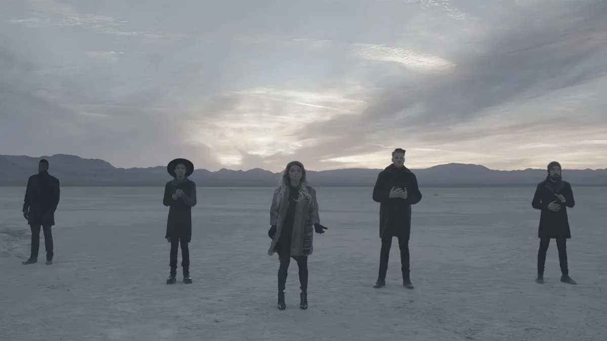 Five people stand in a vast, snowy landscape under a cloudy sky, with mountains in the distance.