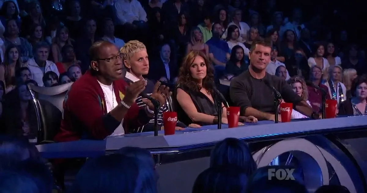 Four judges sit at a table with microphones, clapping, in front of a live audience.