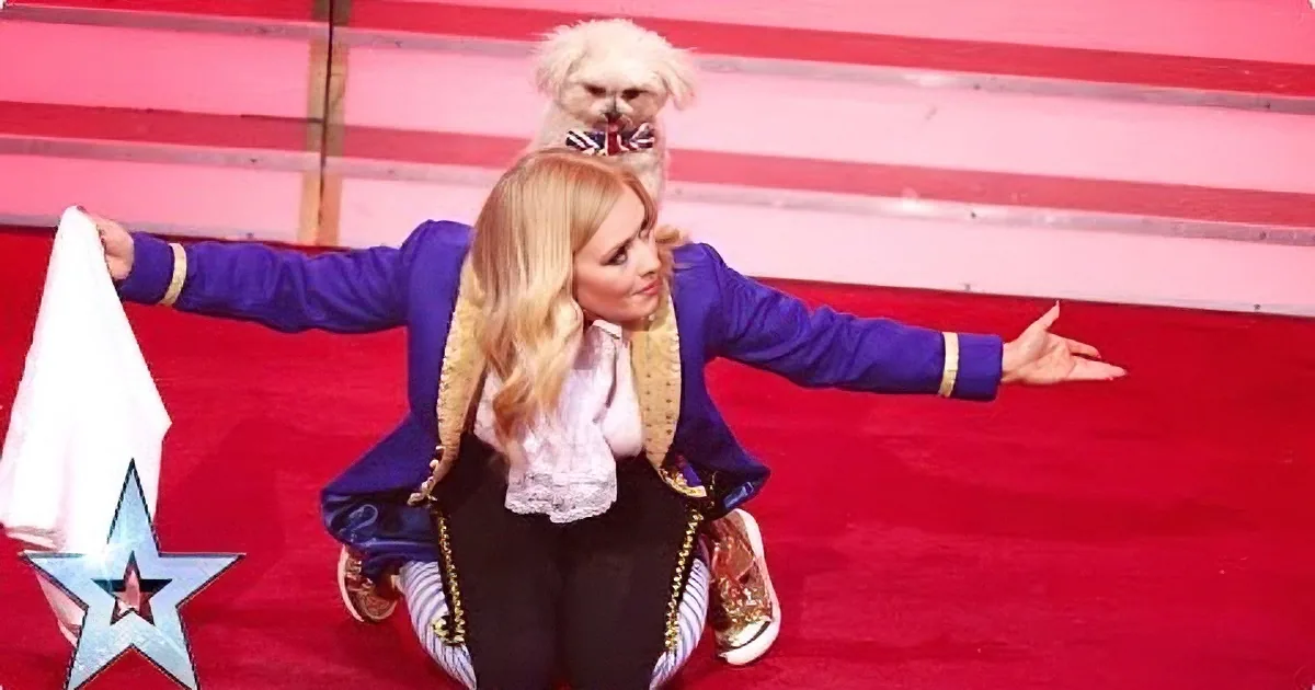 A person in a blue jacket and black pants stands on a red carpet with a small dog on their shoulders.