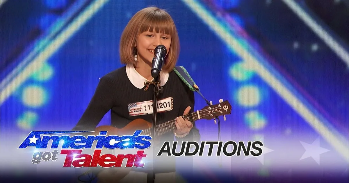 A young girl with short hair plays guitar and sings on stage at America's Got Talent auditions.