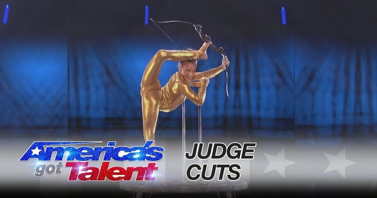 A performer in a gold outfit balances on a pole while holding a bow, showcasing impressive flexibility and strength.