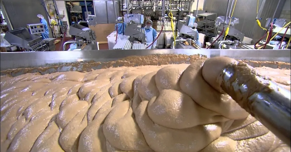 A close-up of a thick, brownish substance being processed in a factory setting with workers in the background.