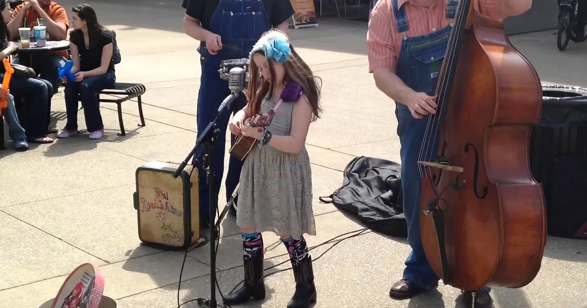 A young girl in a dress and cowboy boots plays a ukulele on a sidewalk, accompanied by a man with a bass.