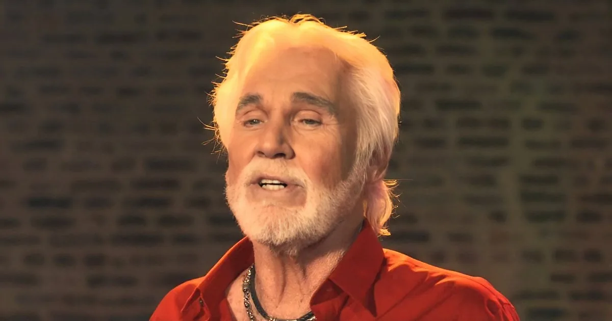 An older man with white hair and beard, wearing a red shirt, is speaking against a textured background.