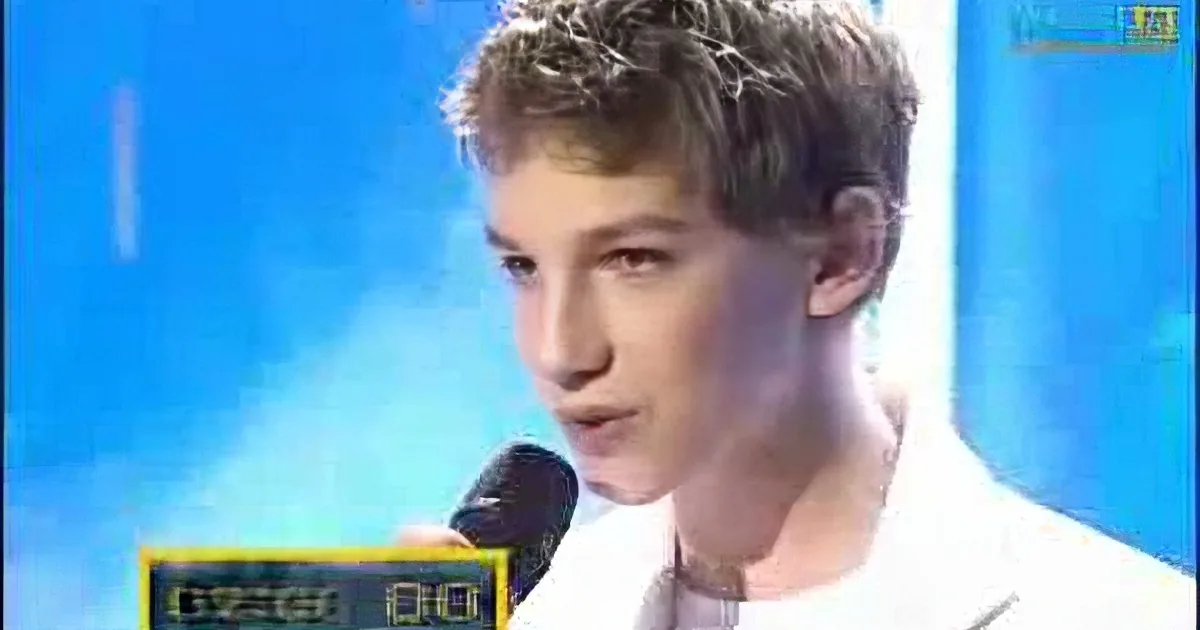 A young person in a white shirt speaks into a microphone against a blue background.