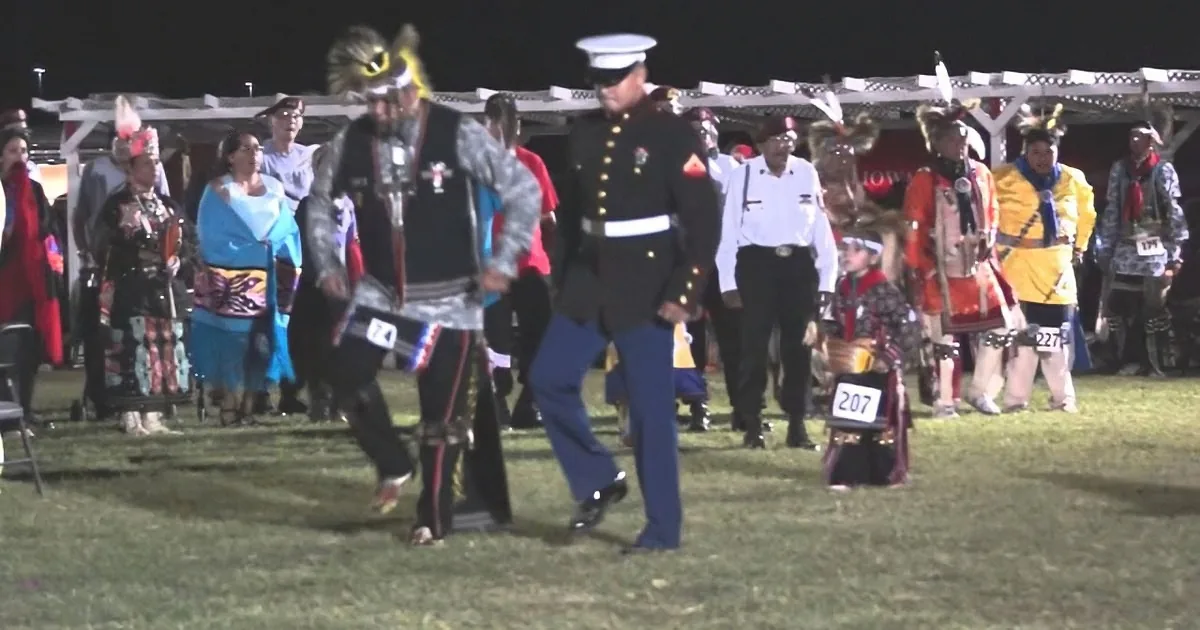 A Marine in uniform dances alongside a Native American in traditional attire at a nighttime event.