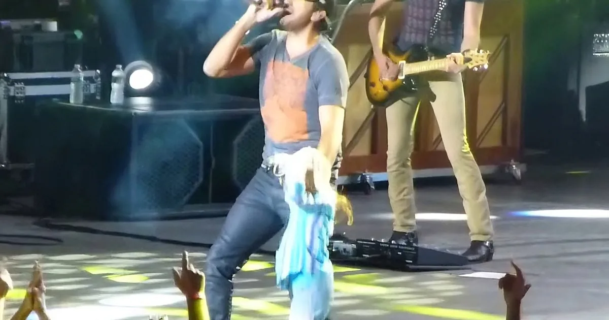 A performer in a blue shirt and jeans is singing on stage, holding a microphone, with a guitarist in the background.