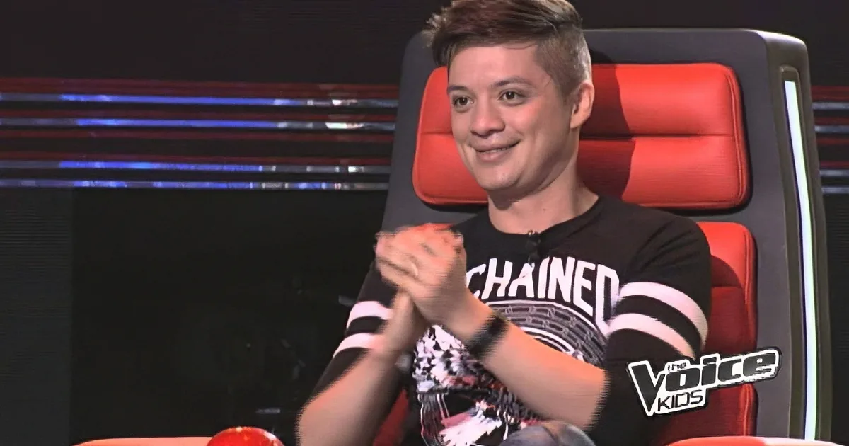 A man in a black shirt with "CHAINED" sits in a red chair, clasping his hands, on a stage with "The Voice Kids" logo.