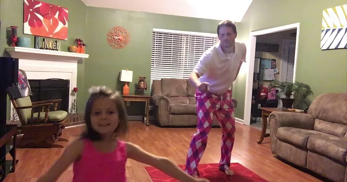 A man in checkered pants dances with a young girl in a pink dress in a cozy living room.
