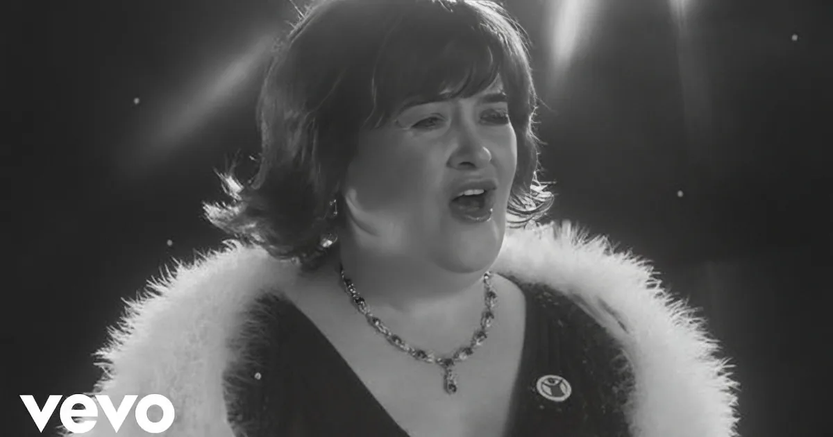 A woman in a fur-trimmed outfit sings passionately, wearing a necklace, with a spotlight effect behind her.
