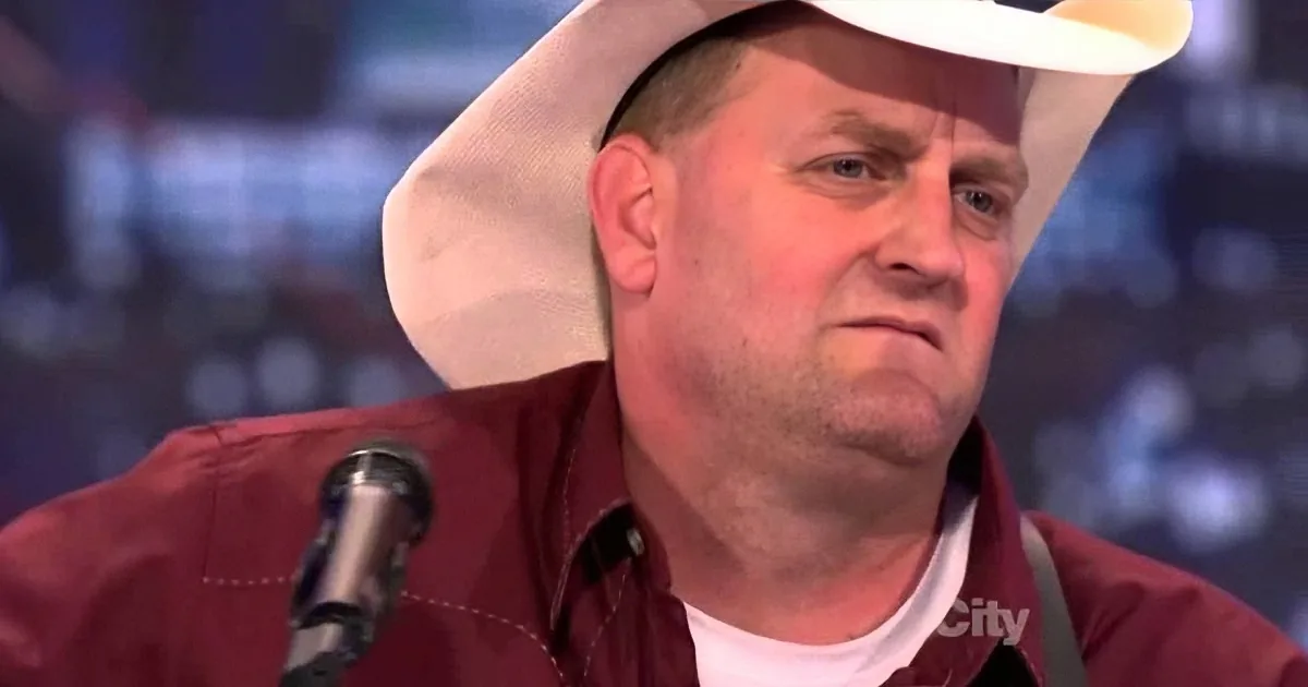 A man wearing a white cowboy hat and a maroon jacket sits in front of a microphone, appearing to be in a studio setting.