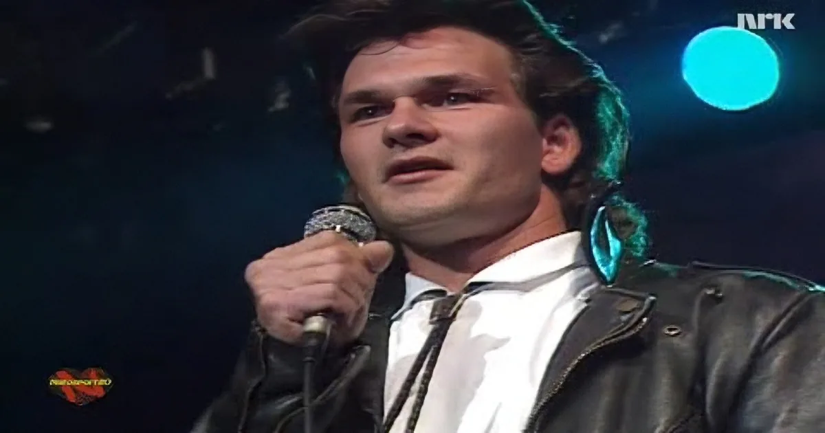 A man in a leather jacket and white shirt sings into a microphone on stage.