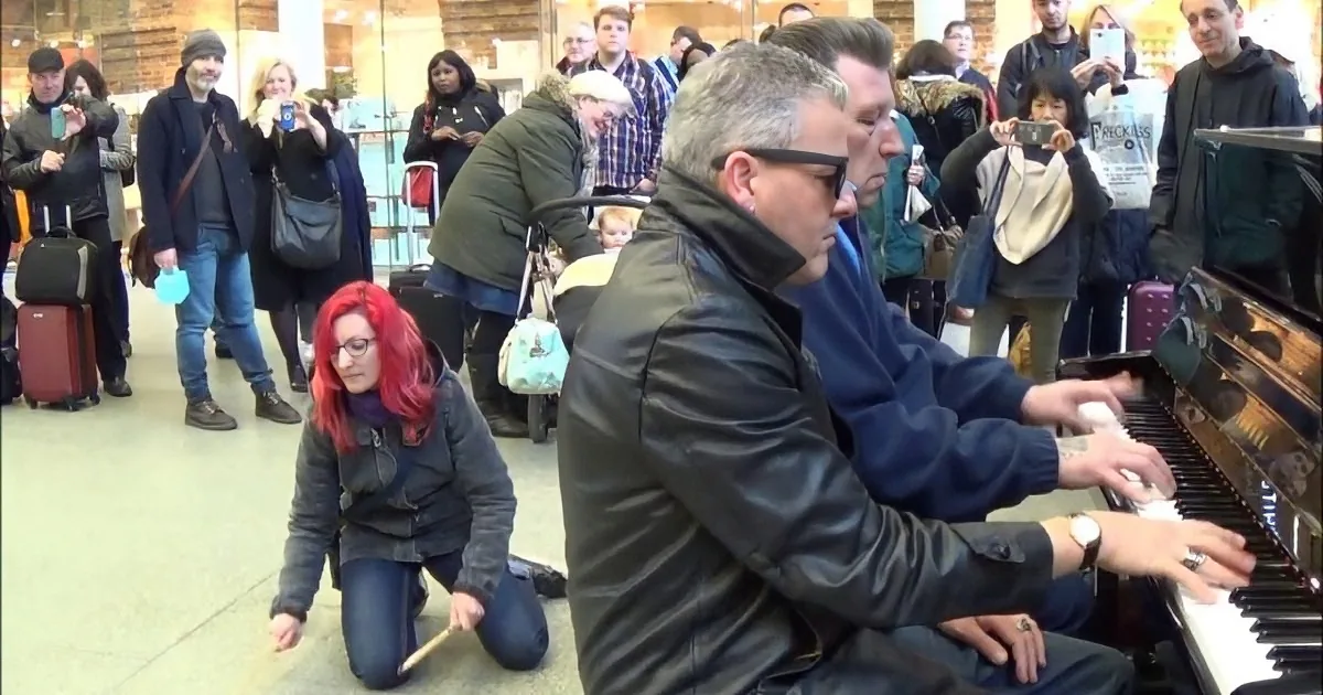 A man in a black jacket plays a piano while a woman kneels beside him, surrounded by onlookers in a busy indoor setting.