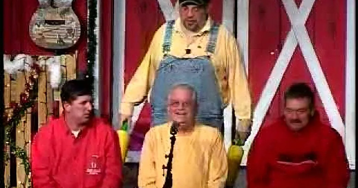 A man in yellow overalls and a yellow shirt stands on a podium, surrounded by three seated individuals in red jackets.
