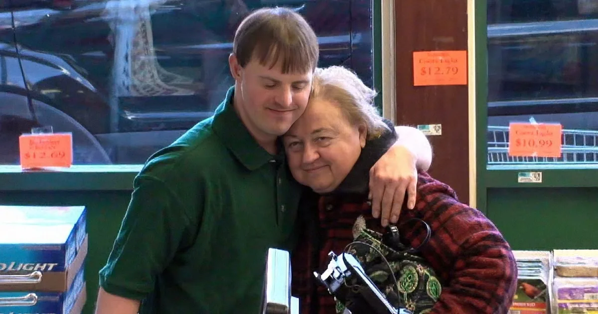 A man in a green shirt hugs an older woman in a plaid shirt at a store checkout.