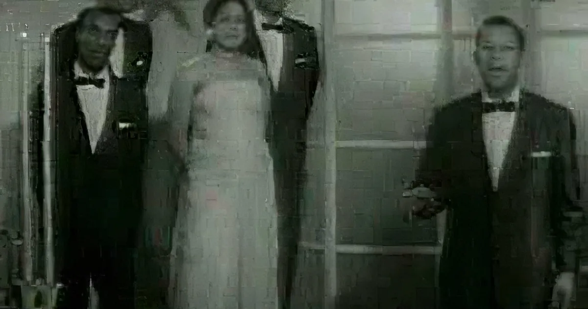 A bride in a white dress stands between two men in tuxedos, smiling.
