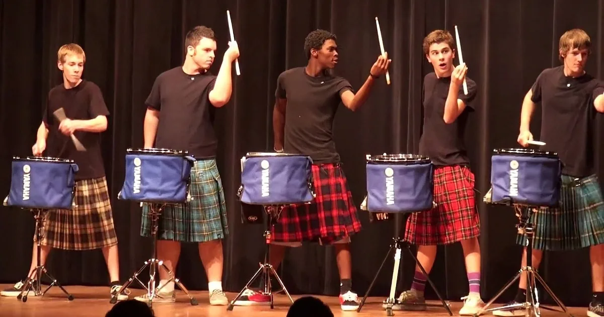 Five young men in kilts play drums on stage, holding drumsticks.