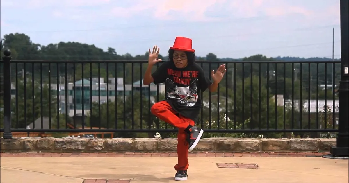 A person in a red hat and black shirt with a cat design dances on a paved area with a scenic background.