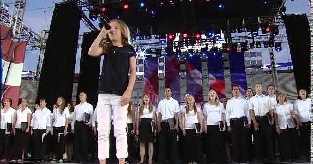 A woman in black and white stands on stage, addressing a choir in white shirts and black pants.