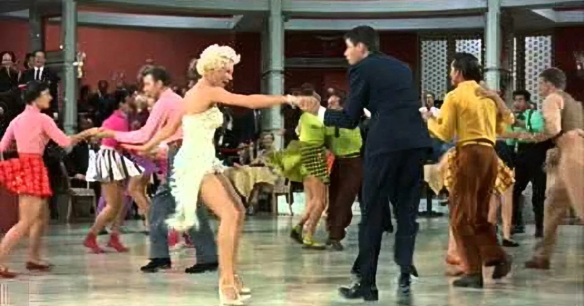 A woman in a white dress dances with a man in a black suit in a lively, crowded dance hall.