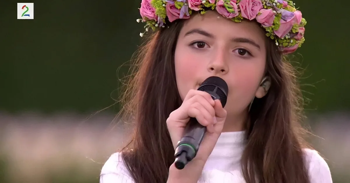 A girl wearing a floral crown sings into a microphone, holding it with both hands.