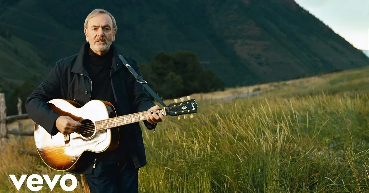 A man in a black jacket holds an acoustic guitar in a grassy field with mountains in the background.