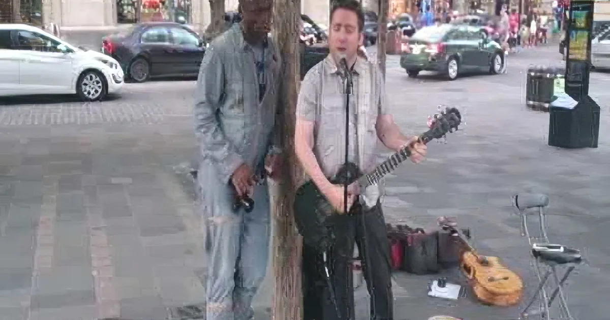 A man plays guitar on a city sidewalk, another man stands nearby, both appear to be busking.