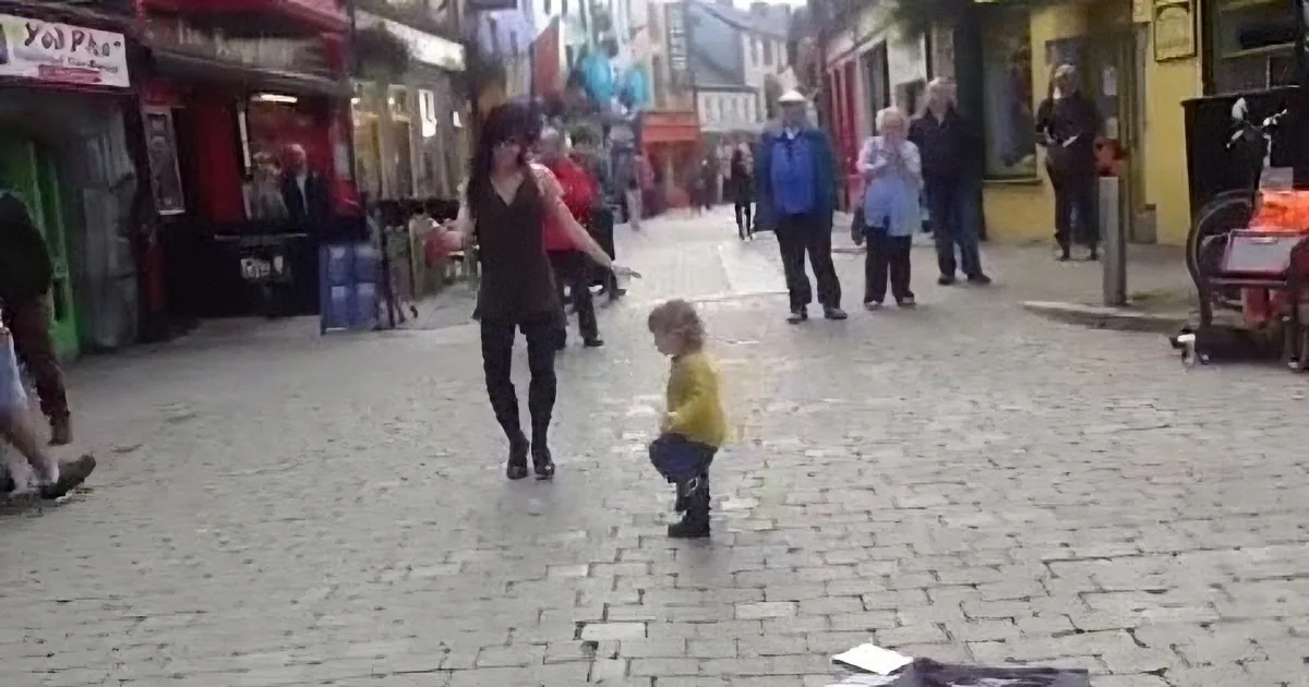 A woman and a child dance in a cobblestone street surrounded by onlookers and shops.
