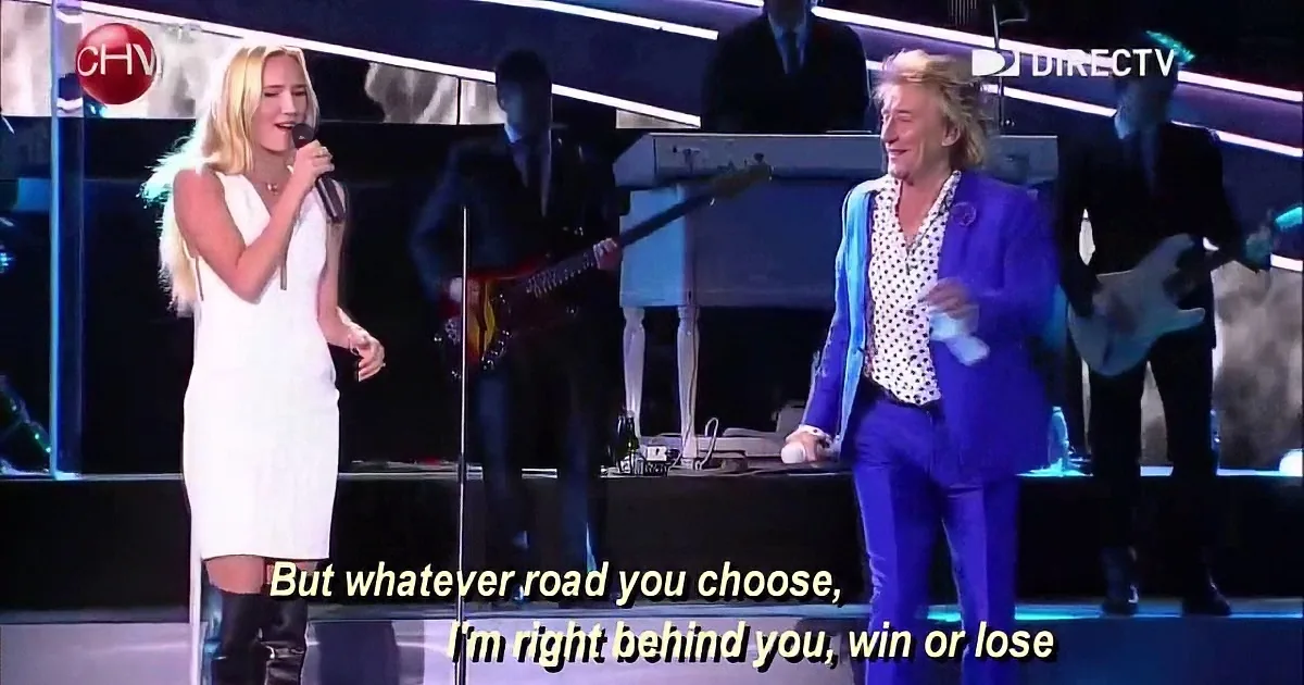 A woman in white sings on stage, accompanied by a man in blue, with a band in the background.