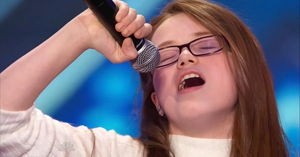 A person with glasses sings passionately into a microphone against a blue background.