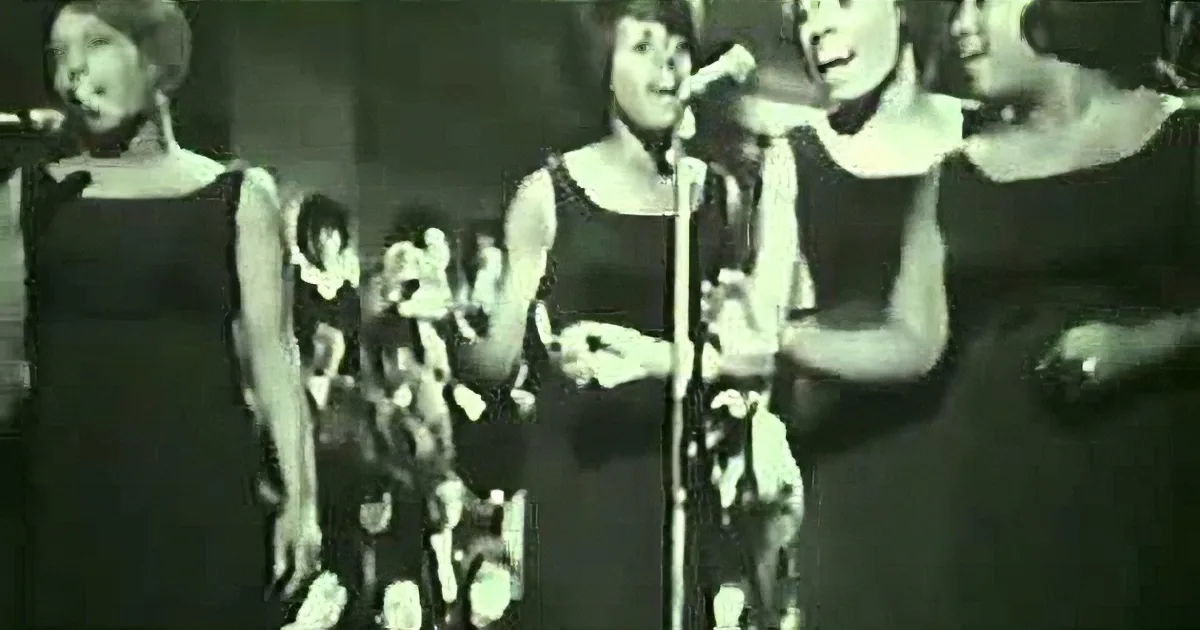 A woman in a black dress sings into a microphone, surrounded by other women in similar attire.