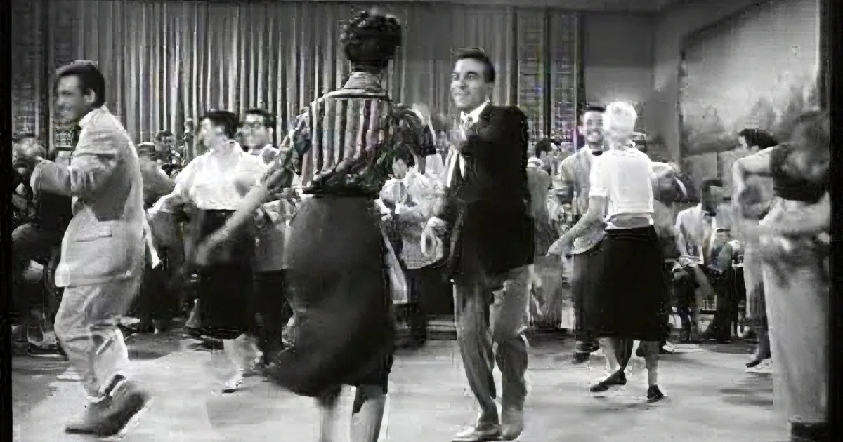 A man in a suit dances with a woman in a striped shirt and skirt in a crowded room.