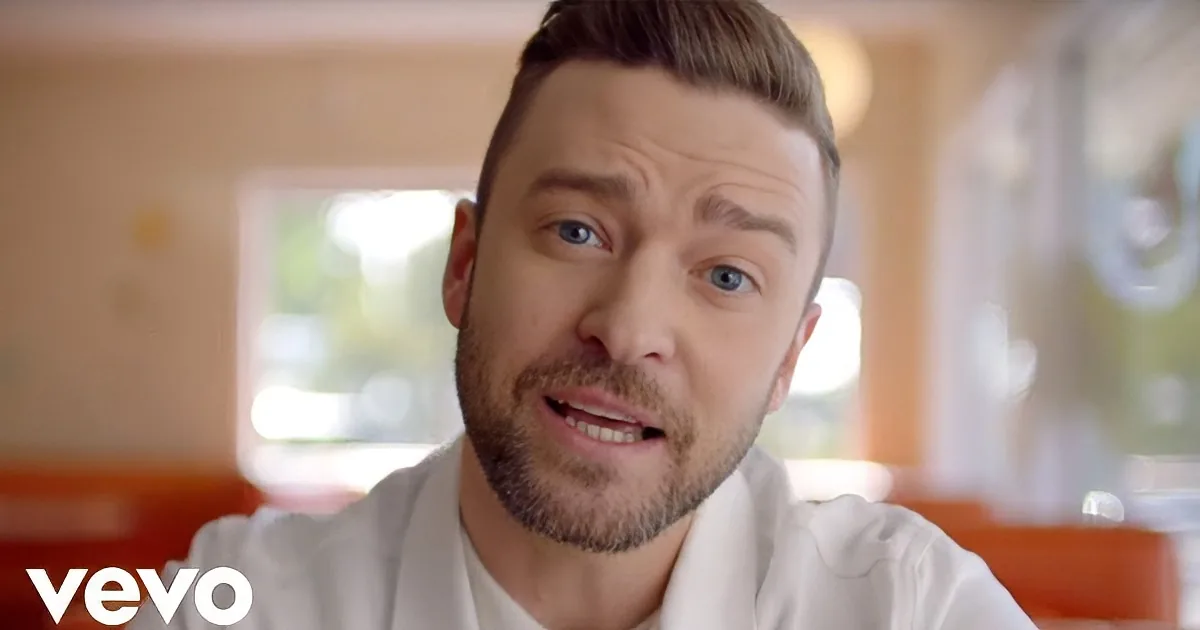 A man with short hair and a beard, wearing a white shirt, is looking directly at the camera with a slight smile.