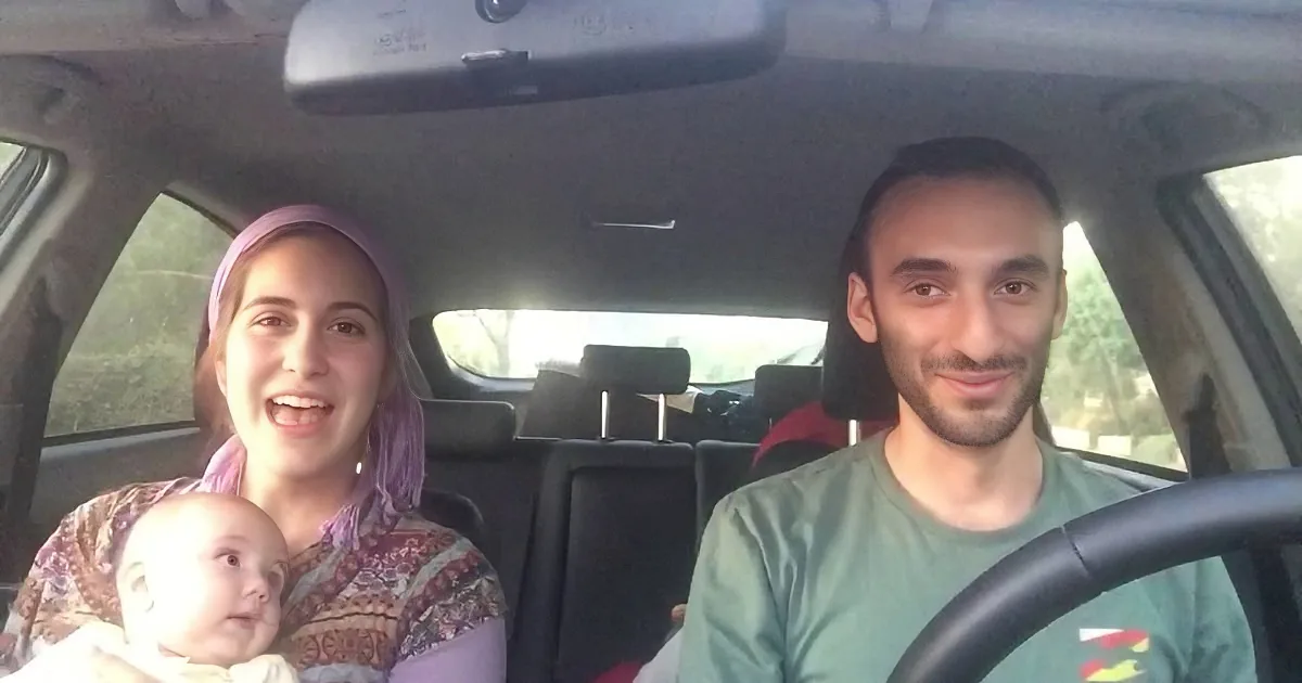 A family of three smiles in a car, with a baby held by the woman in the passenger seat.
