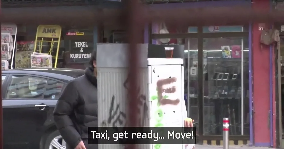 A man in a black jacket stands near a parked car, with a store in the background and a sign saying "Taxi, get ready... Move!"