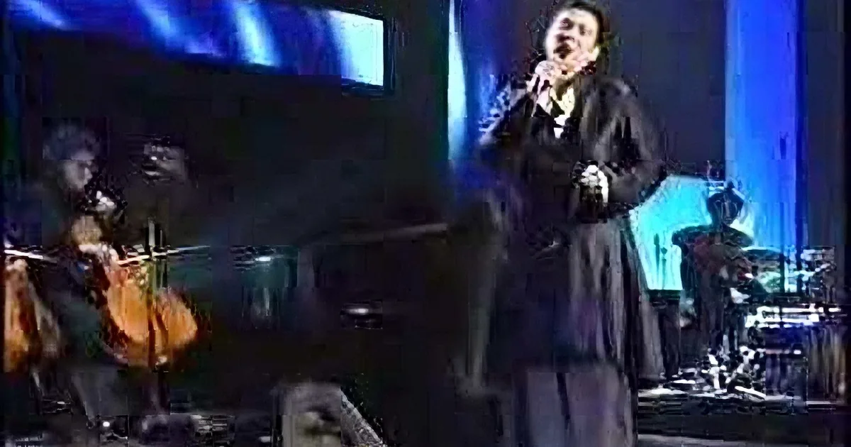 A man in a long coat sings passionately on stage with musicians playing in the background.