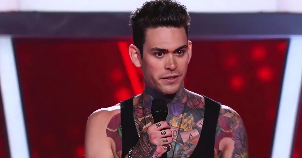 A man with tattoos speaks into a microphone on a stage with red and white lighting.