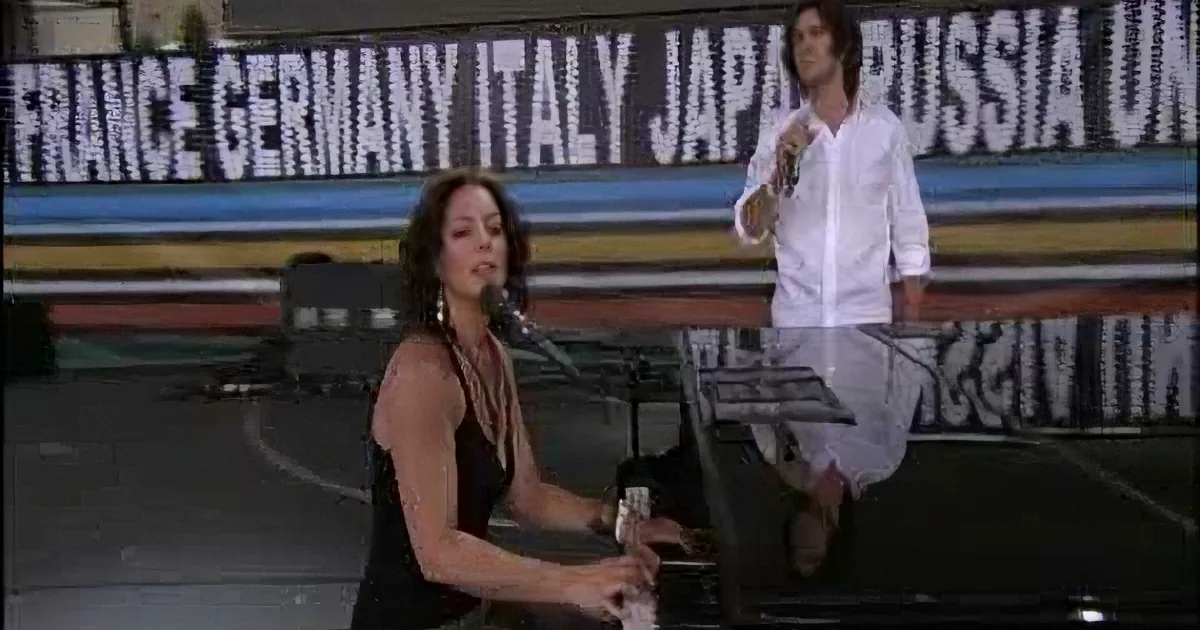 A woman in a black dress sits at a piano, singing into a microphone, with a man in a white shirt standing behind her.