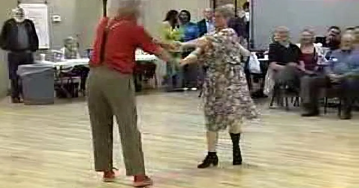 An elderly couple dances in a community center, surrounded by spectators.