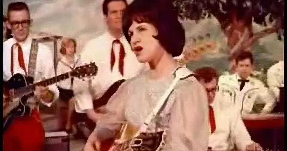 A woman in a white outfit sings passionately while playing a guitar, surrounded by a band in white shirts and ties.