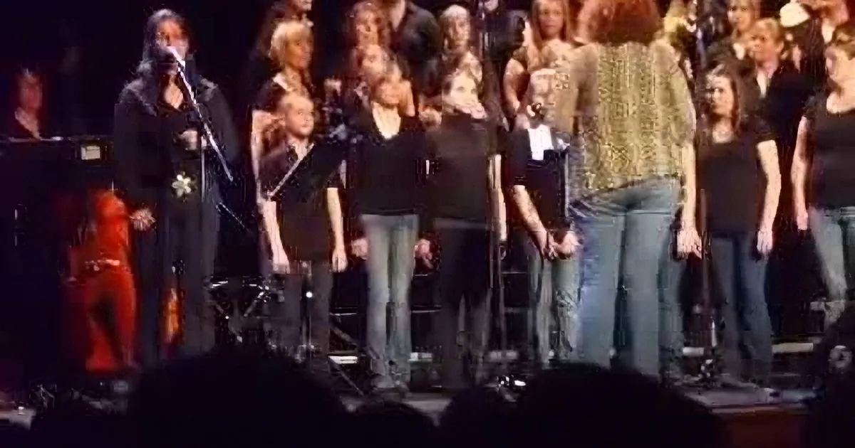 A group of people, including a woman in red, are singing on stage with microphones and a keyboard.