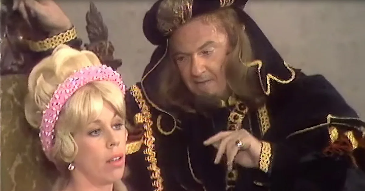 A man in elaborate black and gold attire is gesturing while seated next to a woman in a pink beaded headband.