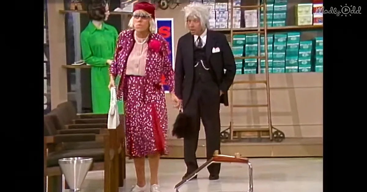 A woman in a red floral dress and hat stands next to a man in a suit and white wig in a store setting.
